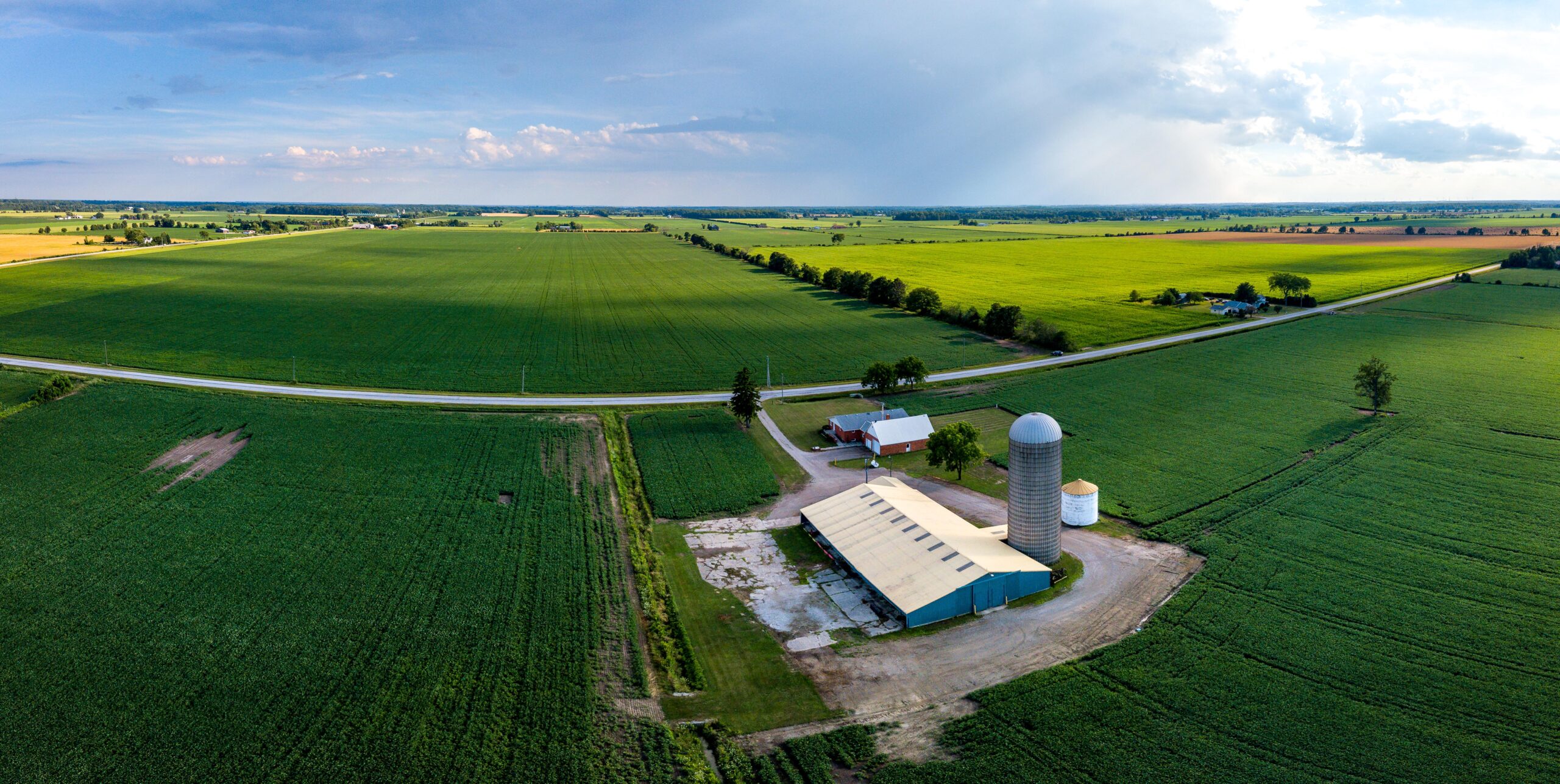 Aerial view of a lush, expansive farmland with neat green fields under a clear sky. A farmstead with a red barn, silo, and large agricultural buildings sits near a winding road, embodying the thriving agricultural landscape of Ontario.